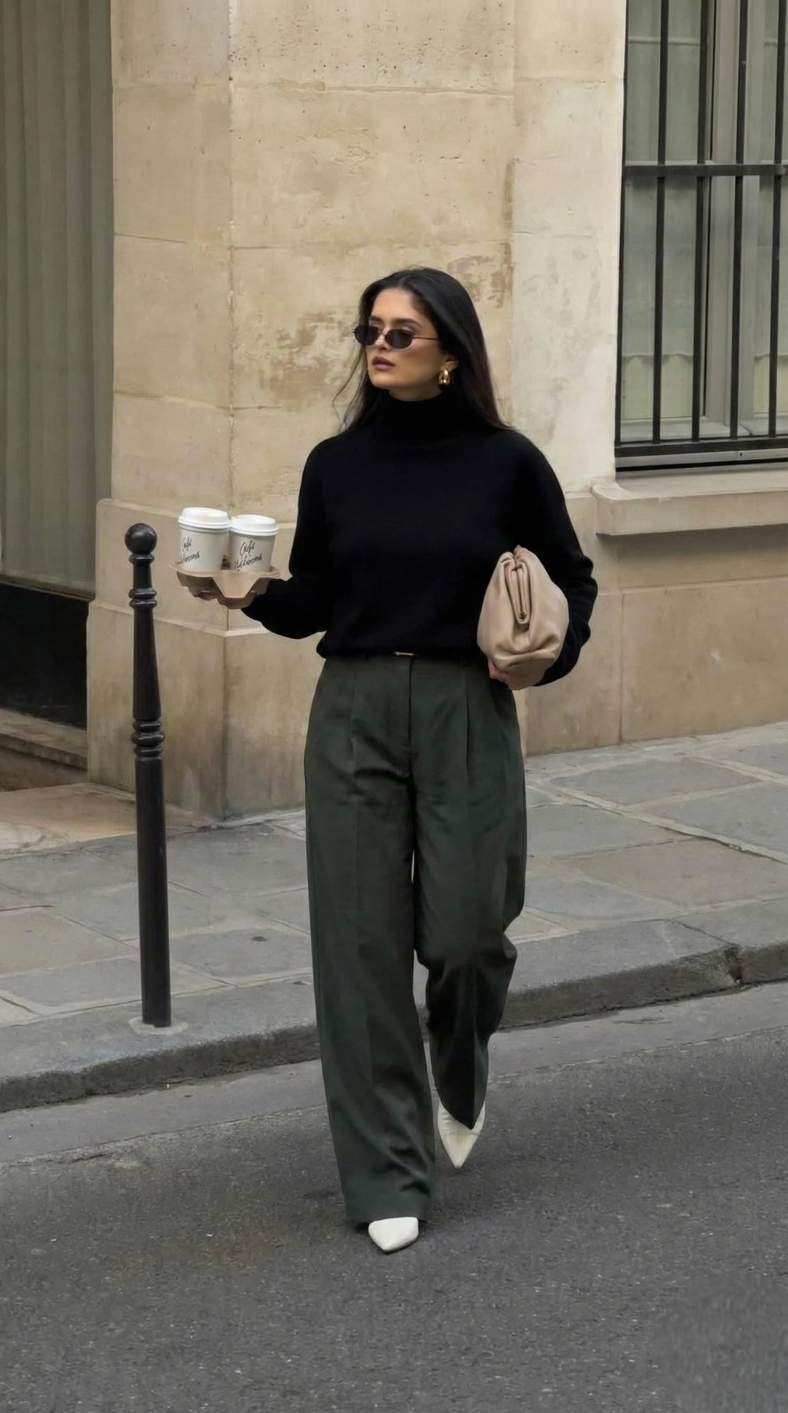 Pull col roulé noir, pantalon large vert olive, sac pochette beige et cafés en main