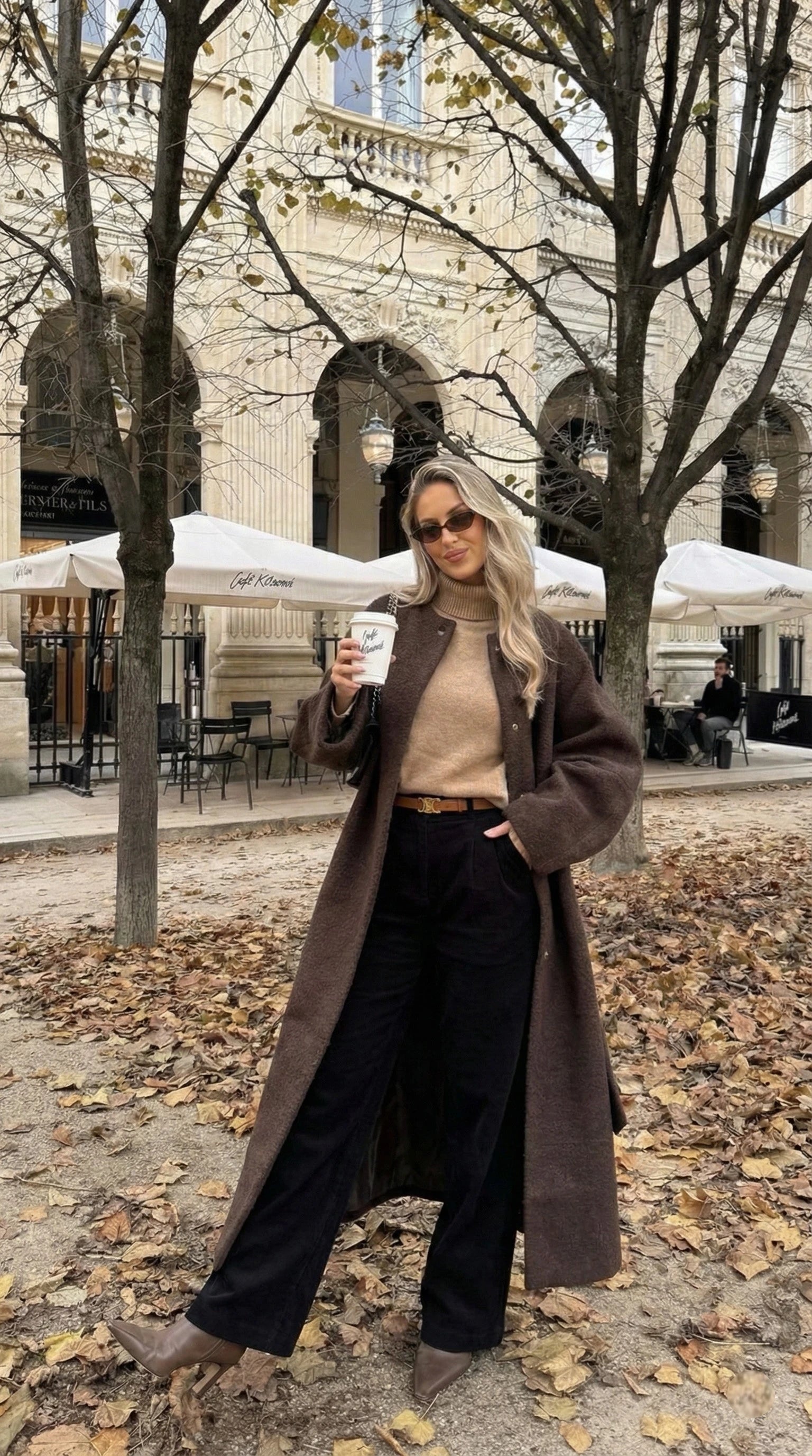Manteau long marron, pull col roulé beige, pantalon noir et café à la main